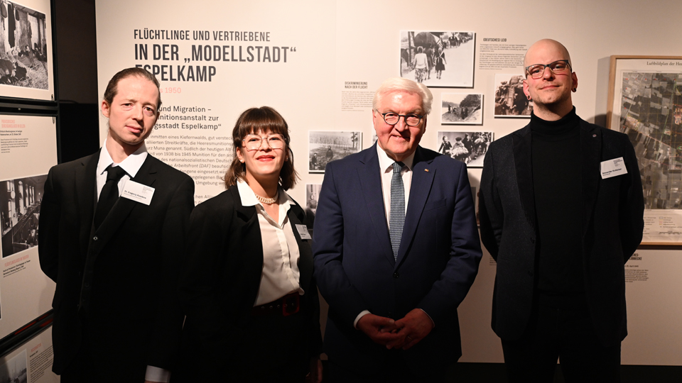 Drei Männer und eine Frau stehen nebeneinander vor einem Exponat der Ausstellung und lächeln in die Kamera.