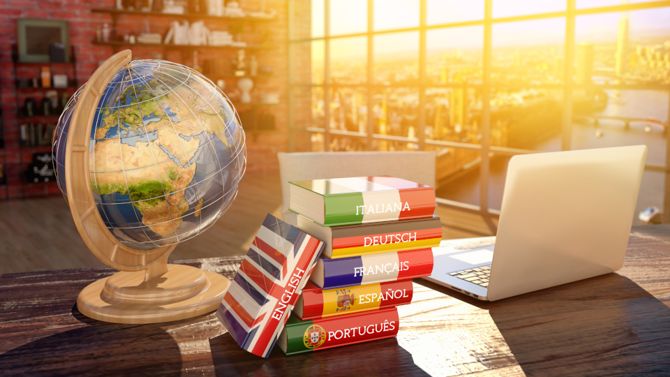 Auf einem Schreibtisch stehen von links nach rechts: ein Globus, ein Stapel Bücher mit Flaggen als Mantel, auf den Buchrücken steht jeweils eine Sprach: Englisch, Italienisch, Deutsch, Französisch, Spanisch und Portugiesisch; rechts daneben ein Laptop.