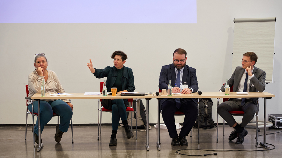 Zwei Frauen und zwei Männer sitzen an Tischen als Teilnehmende an einer Podiumsdiskussion