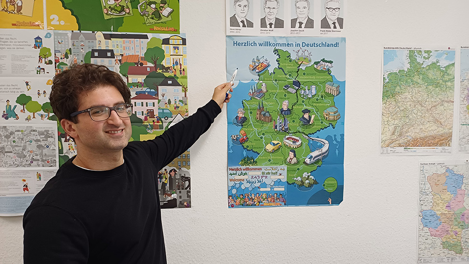 Ein Mann steht vor einer Wand. Er zeigt auf ein Plakat, das an der Wand hängt. (Quelle: Juliane Quägwer | Johanniter-Unfall-Hilfe e. V.) Ein Mann steht vor einer Wand. Er zeigt auf ein Plakat, das an der Wand hängt.