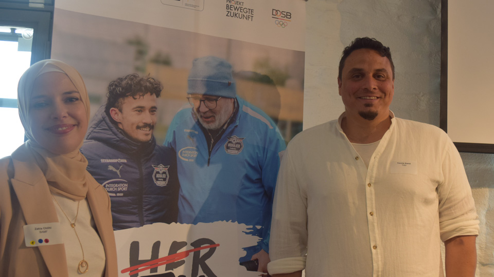 Zwei Personen stehen vor einem Banner. (Quelle: BAMF | Anke Lübbert) Zwei Personen stehen vor einem Banner.