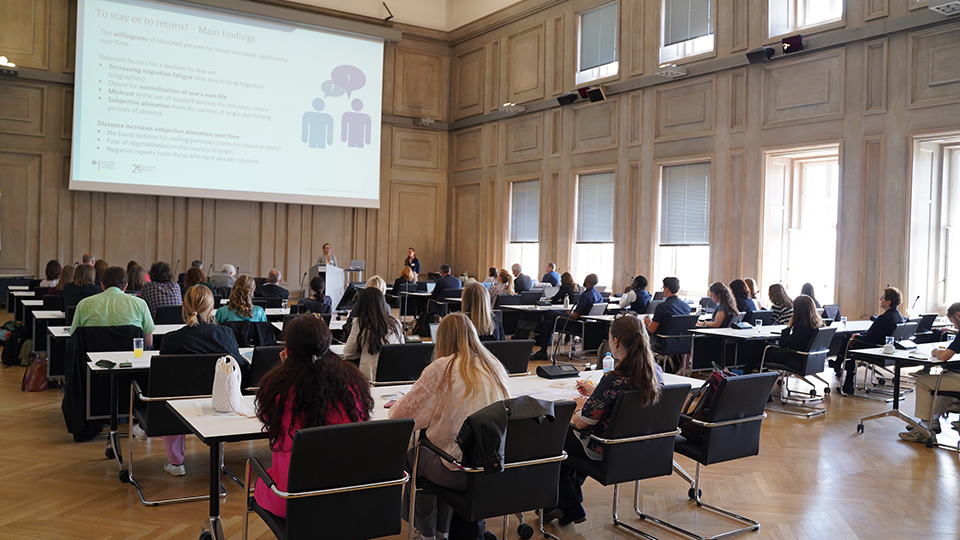 Viele Personen sitzen in einem großen Konferenzraum und schauen auf die Rednerin am Rednerpult und die Präsentation vor ihnen. (Quelle: BAMF) Viele Personen sitzen in einem großen Konferenzraum und schauen auf die Rednerin am Rednerpult und die Präsentation vor ihnen.