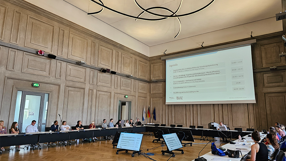 Zahlreiche Teilnehmende sitzen an Tischen im großen Konferenzsaal des Bundesamtes. Auf eine Leinwand ist eine Präsentation mit der Agenda der Veranstaltung projiziert.  (Quelle: BAMF) Zahlreiche Teilnehmende sitzen an Tischen im großen Konferenzsaal des Bundesamtes. Auf eine Leinwand ist eine Präsentation mit der Agenda der Veranstaltung projiziert.