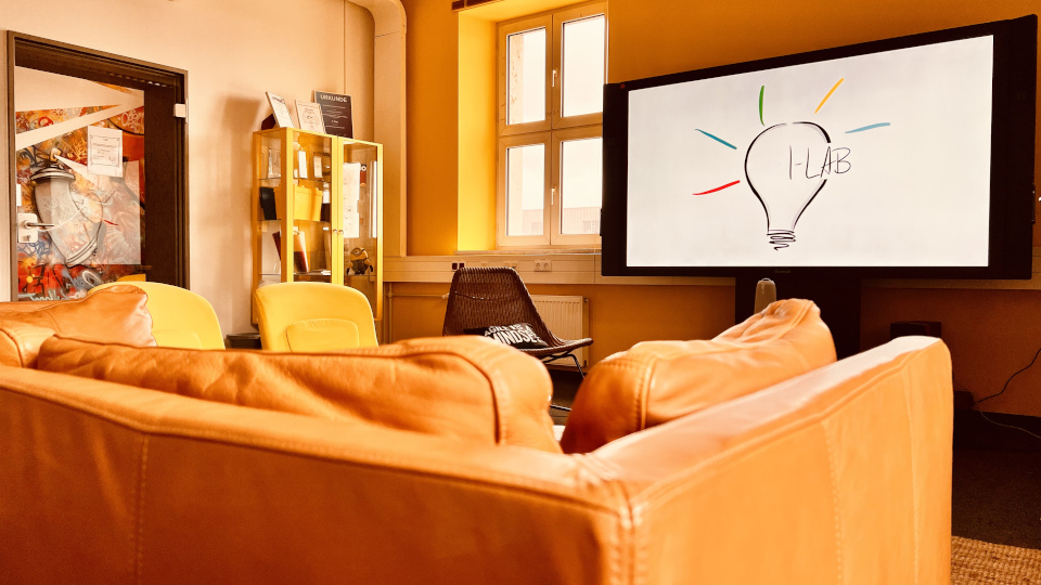 In einem Raum stehen ein Sofa und mehrere Sessel vor einem großen Bildschirm, der eine gezeichnete Glühbirne und den Schriftzug I-LAB zeigt.
