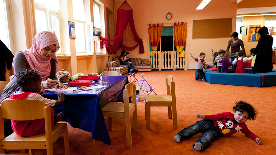 Eine Frau sitzt mit einem Kind an einem Tischen und malt. Daneben spielt ein Kind auf dem Boden. Im Hintergrund spielt eine Gruppe Kinder mit zwei Erwachsenen. (Quelle: Europäische Union) Eine Frau sitzt mit einem Kind an einem Tischen und malt. Daneben spielt ein Kind auf dem Boden. Im Hintergrund spielt eine Gruppe Kinder mit zwei Erwachsenen.