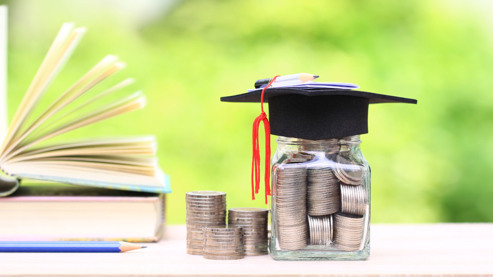 Auf einem Tisch liegen von links nach rechts: Bücher, ein Stift, Geld und ein mit Geld gefülltes Glas. Auf dem Glas liegt ein Doktorhut. Auf dem Hut liegen ein Stift und ein gefalteter Zettel. (Quelle: Monthira / stock.adobe.com) Auf einem Tisch liegen von links nach rechts: Bücher, ein Stift, Geld und ein mit Geld gefülltes Glas. Auf dem Glas liegt ein Doktorhut. Auf dem Hut liegen ein Stift und ein gefalteter Zettel.