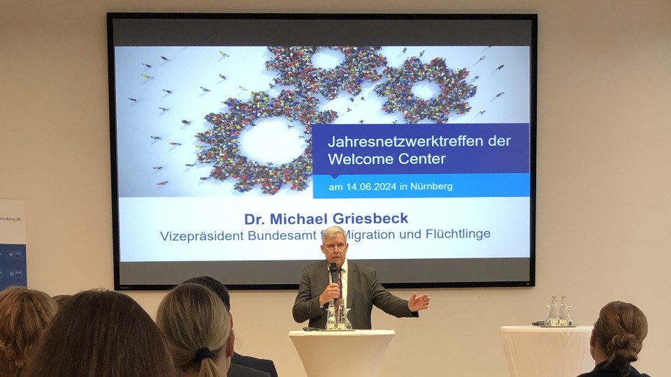 Ein Mann steht an einem Tisch und spricht in ein Mikrofon. Seinen linken Arm hat er leicht ausgestreckt. Hinter ihm hängt ein großer Bildschirm mit einer Präsentation. Vor ihm sitzen einige Zuhörende. (Quelle: BAMF) Ein Mann steht an einem Tisch und spricht in ein Mikrofon. Seinen linken Arm hat er leicht ausgestreckt. Hinter ihm hängt ein großer Bildschirm mit einer Präsentation. Vor ihm sitzen einige Zuhörende.